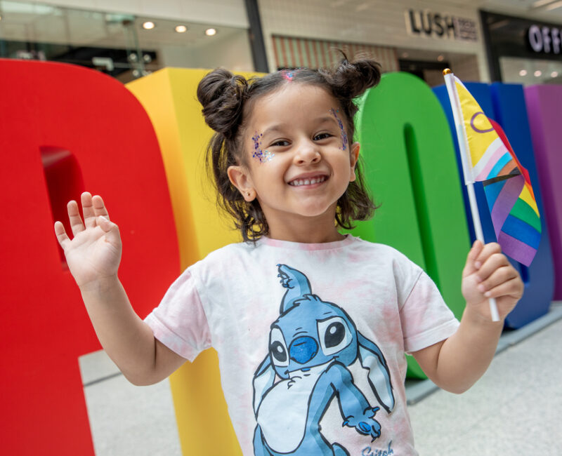 Pride 2025 Little girl waving a LGBTQ+ flag in front of giant multi-coloured "PROUD" letters