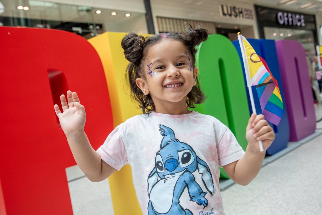 Pride 2025 Little girl waving a LGBTQ+ flag in front of giant multi-coloured "PROUD" letters